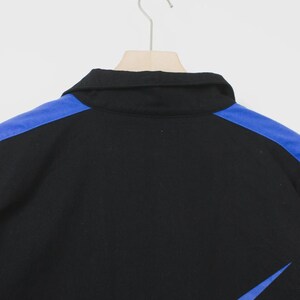 Op de afbeelding: Zwart overhemd met kraag, blauwe schouderaccenten en een blauw schuin ontwerp aan de rechteronderkant. Het overhemd hangt aan een houten hanger tegen een witte achtergrond. De kraag van het overhemd is zwart.