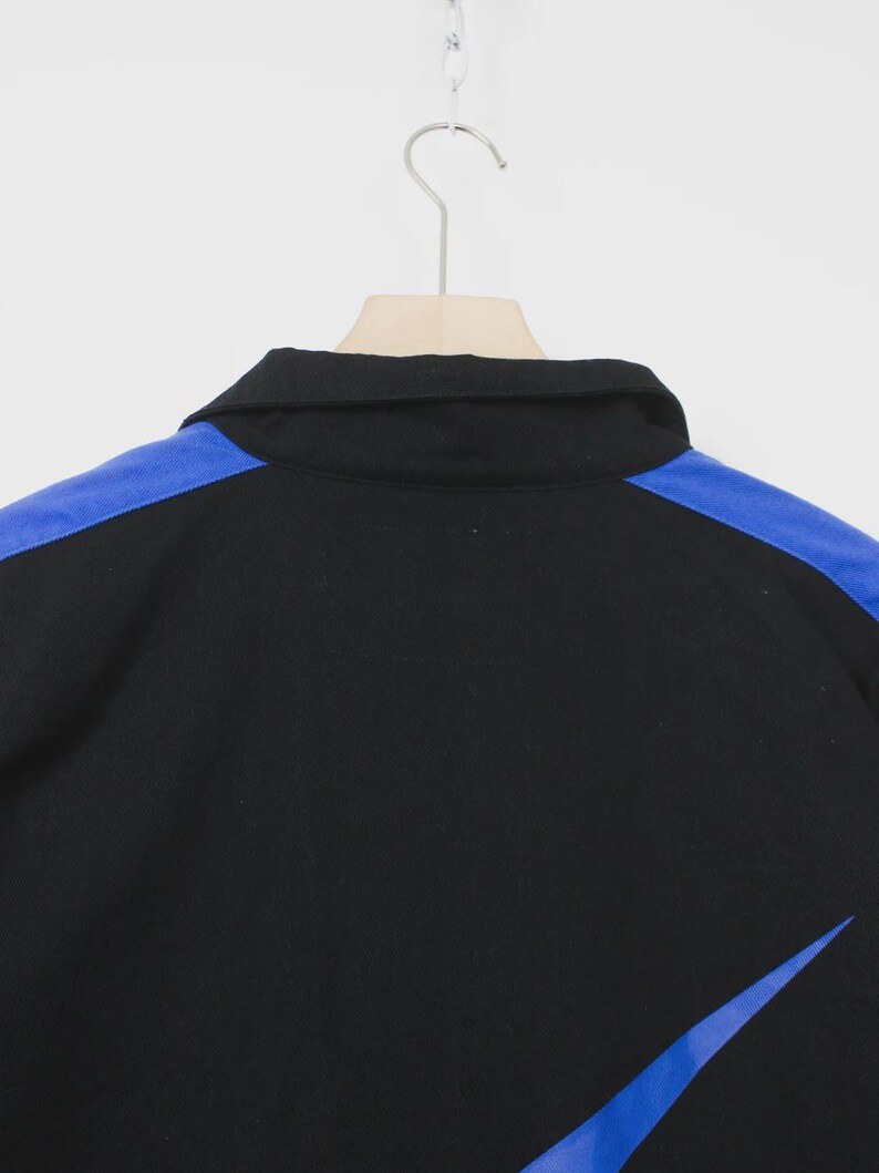 Op de afbeelding: Zwart overhemd met kraag, blauwe schouderaccenten en een blauw schuin ontwerp aan de rechteronderkant. Het overhemd hangt aan een houten hanger tegen een witte achtergrond. De kraag van het overhemd is zwart.