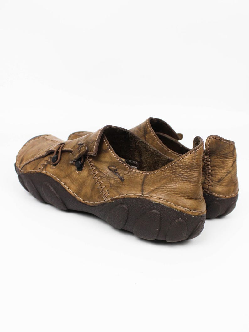 K&ouml;nnte beinhalten: Ein Paar hellbraune Lederschuhe mit Used-Optik, sichtbaren Kontrastn&auml;hten, einer dunkelbraunen, strukturierten Gummisohle und einem Slipper-Design mit einem kleinen seitlichen Riemendetail. Der Markenname Clarks ist seitlich eingepr&auml;gt.