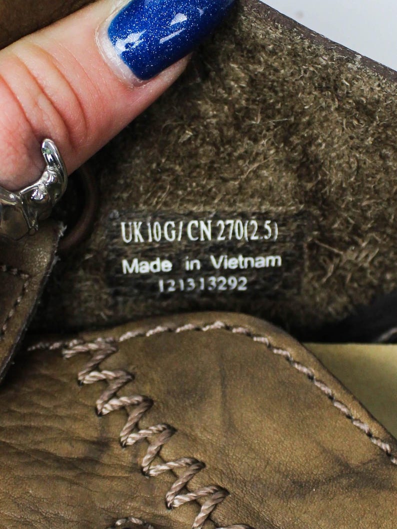 K&ouml;nnte beinhalten: Das Innere eines braunen Lederschuhs mit einem schwarzen Etikett, das den wei&szlig;en Text UK 10G / CN 270(2.5), Made in Vietnam und die Nummer 121313292 tr&auml;gt. Der Schuh hat sichtbare beige N&auml;hte an der Lederkante und ein strukturiertes Innenfutter.