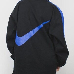 Op de afbeelding: Zwarte jas met een groot blauw Nike swoosh-logo op de rug en een blauwe streep over de rechter mouw. De jas heeft een kraag en een ritssluiting. De jas is gemaakt van duurzaam materiaal.