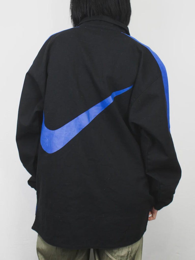 Op de afbeelding: Zwarte jas met een groot blauw Nike swoosh-logo op de rug en een blauwe streep over de rechter mouw. De jas heeft een kraag en een ritssluiting. De jas is gemaakt van duurzaam materiaal.