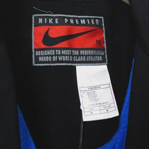 Op de afbeelding: Zwarte Nike Premier jas met een rood en zwart logo patch. De patch leest "NIKE PREMIER" en "DESIGNED TO MEET THE PERFORMANCE NEEDS OF WORLD CLASS ATHLETES". Een wit label met tekst is bevestigd.