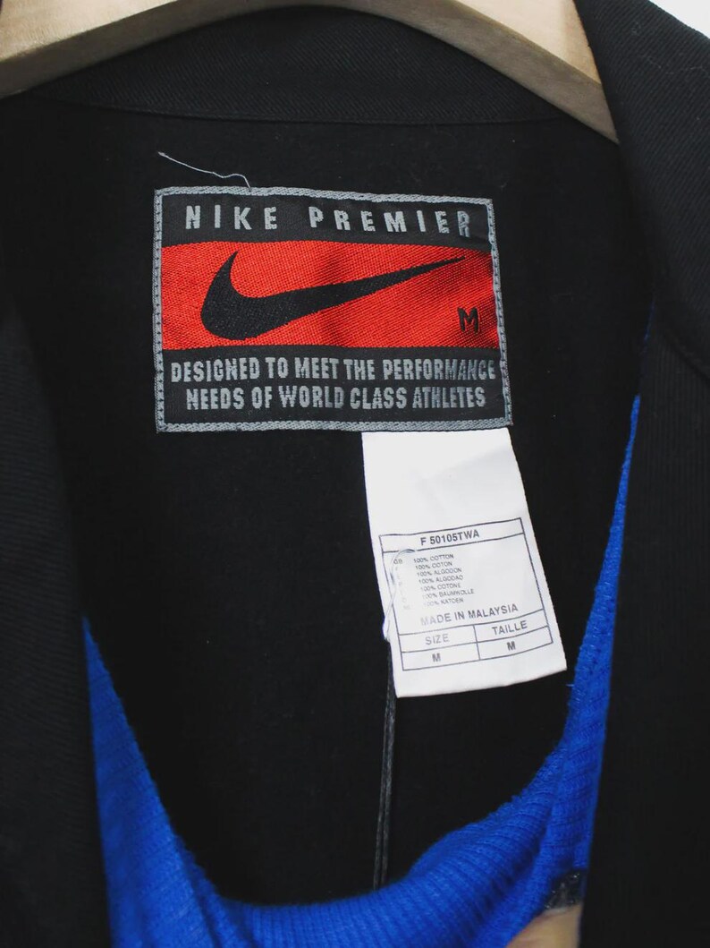 Op de afbeelding: Zwarte Nike Premier jas met een rood en zwart logo patch. De patch leest "NIKE PREMIER" en "DESIGNED TO MEET THE PERFORMANCE NEEDS OF WORLD CLASS ATHLETES". Een wit label met tekst is bevestigd.