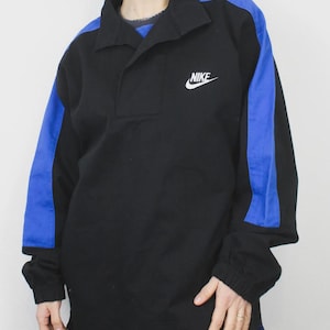 Op de afbeelding: Zwart shirt met lange mouwen en blauwe schouder- en mouwaccenten. Voorzien van een kraag en een klein wit Nike-logo. De mouwen hebben elastische manchetten.