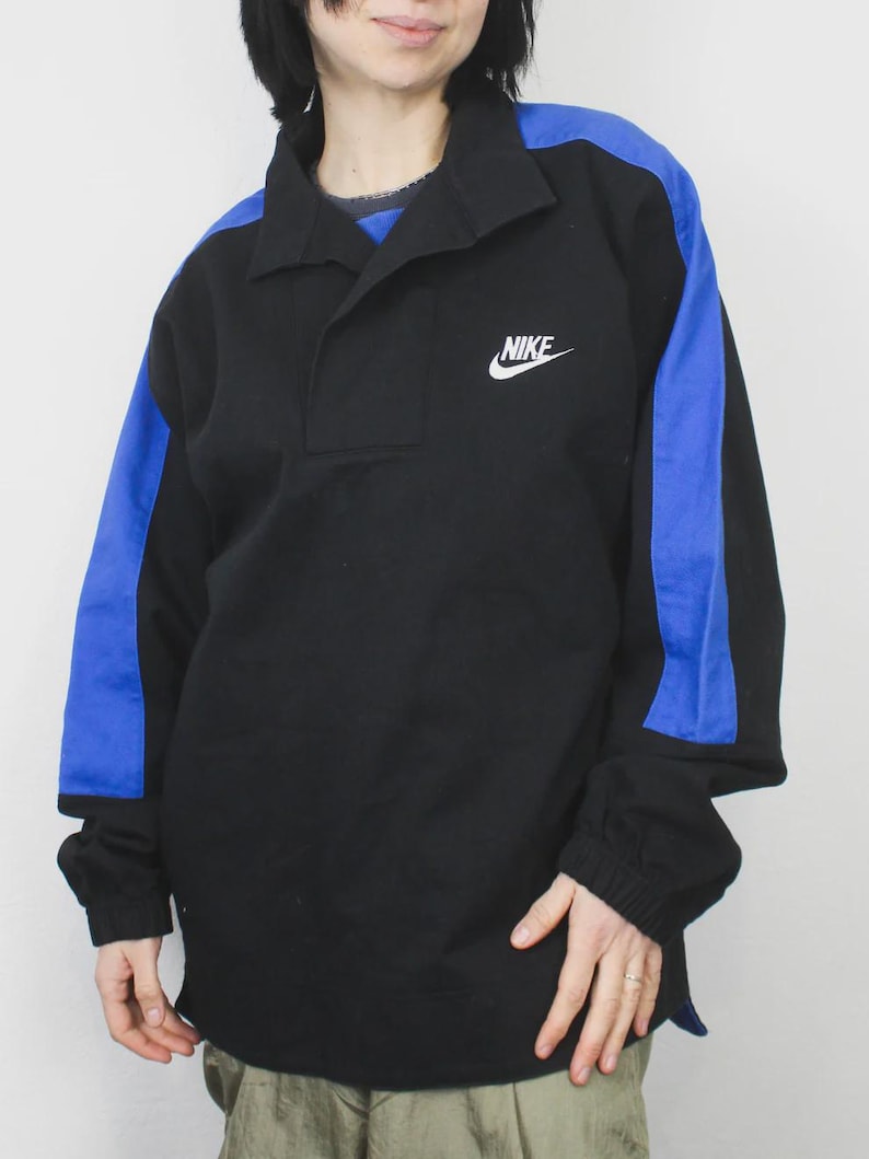 Op de afbeelding: Zwart shirt met lange mouwen en blauwe schouder- en mouwaccenten. Voorzien van een kraag en een klein wit Nike-logo. De mouwen hebben elastische manchetten.