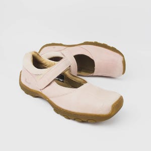 Może przedstawiać: Para jasnoróżowych butów w stylu Mary Jane z paskiem i brązową podeszwą. Buty wykonane są z miękkiego materiału i mają zaokrąglony nosek. Buty mają około 23 cm długości.