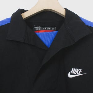 Op de afbeelding: Zwart overhemd met kraag, blauwe schouderaccenten en een wit Nike-logo. Het overhemd heeft een blauw-rode geribbelde kraag en de tekst "NIKE PREMIER" op het label.