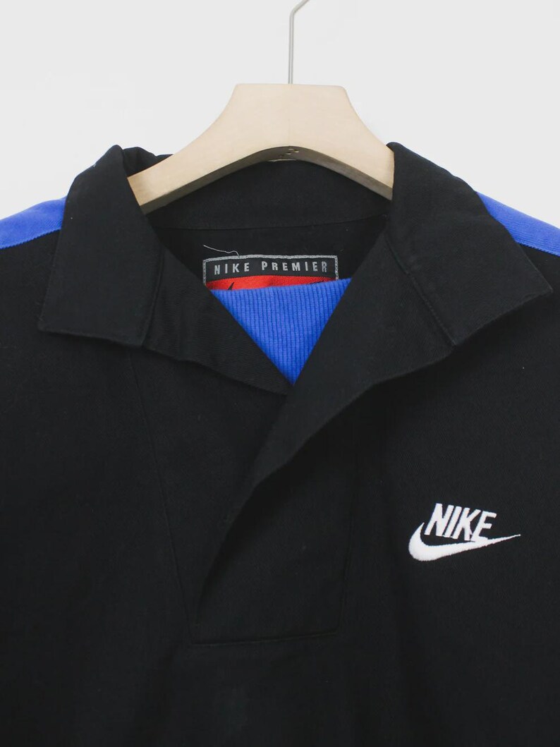 Op de afbeelding: Zwart overhemd met kraag, blauwe schouderaccenten en een wit Nike-logo. Het overhemd heeft een blauw-rode geribbelde kraag en de tekst "NIKE PREMIER" op het label.