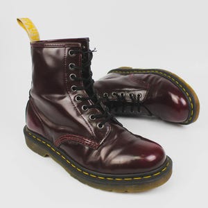 Puede incluir: Par de botas de cuero burdeos con cordones negros y pespuntes amarillos. Las botas tienen un acabado brillante y una lengüeta amarilla en la parte superior. Las botas son de estilo clásico.