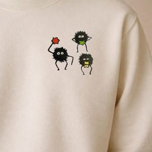May include: Cream-colored sweatshirt featuring a whimsical design of three cartoonish, black, furry creatures with large eyes. One holds a red star, another has a green leaf, and the third has a yellow bow tie. The sweatshirt has a crew neck.