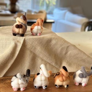 Peut inclure: Six figurines de chat en laine feutrée, chacune avec une couleur et un motif différents, sont disposées en rangée sur une surface en bois. Les chats sont tous tournés dos à la caméra, la queue enroulée sur le dos.