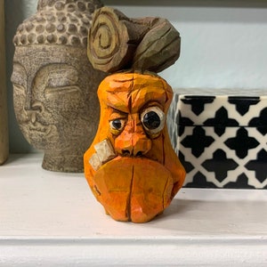 May include: A hand-carved wooden figurine of a grumpy-looking face with a bandage over its eye. The figurine is painted orange and has a log on top of its head.