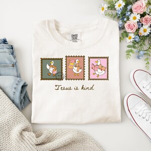 Puede incluir: Camiseta color crema con tres diseños de sellos postales con patos de dibujos animados. Cada pato lleva una bufanda y lleva objetos como un libro o paquetes. El texto "Jesus is kind" está impreso debajo. La camiseta es de algodón.