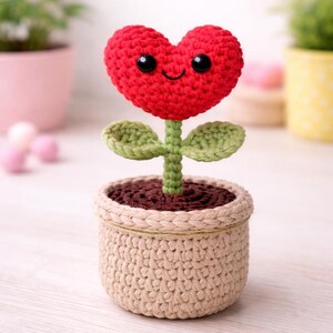 Peut inclure: Une fleur crochetée en forme de cœur rouge souriant, avec des yeux noirs et une tige et des feuilles vertes. Elle est plantée dans un pot beige crocheté rempli de fil marron. Un objet décoratif fait à la main.