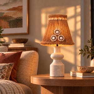 May include: A table lamp with a smooth, off-white, tiered ceramic base and a textured, tan, fringed lampshade. The shade is decorated with three circular, white and tan beaded accents arranged in a triangular pattern near the center.