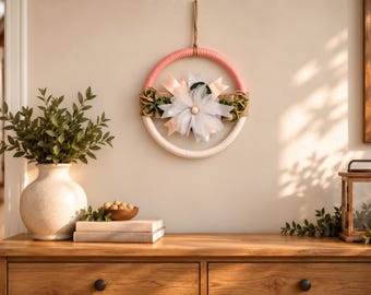 May include: A circular wall hanging featuring a rope-wrapped frame in coral and white. The center is decorated with a layered bow made of white tulle, peach-colored ribbon, and burlap fabric, accented with a small wooden bead at the very center.