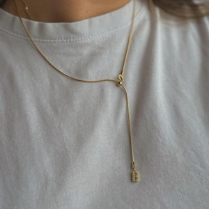 Könnte beinhalten: Eine zarte, goldfarbene Halskette mit Y-förmigem Design. Die Halskette hat eine schlanke Kette und einen kleinen, quadratischen Anhänger am Ende des vertikalen Tropfens. Die Halskette wird auf einem weißen T-Shirt getragen.