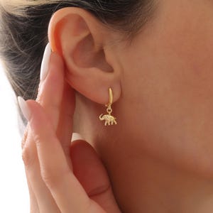 Könnte beinhalten: Ein goldener Creolen-Ohrring mit einem kleinen, hängenden Elefanten-Anhänger. Der Ohrring ist am Ohrläppchen befestigt und zeigt eine polierte metallische Oberfläche sowie eine detaillierte Miniatur-Elefantenfigur, die am Ring hängt.