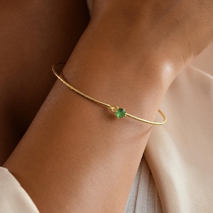 May include: A delicate gold-toned bracelet with a small, round, green gemstone at its center. The bracelet has a thin, hammered texture and is worn on a wrist. The background is a soft, neutral color, highlighting the jewelry.