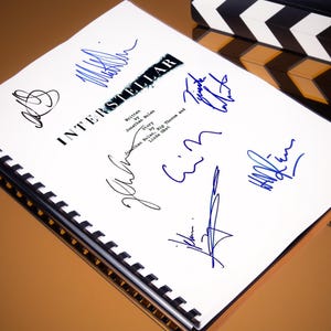 Pode incluir: Um roteiro branco com o título "INTERSTELLAR" escrito em preto. O roteiro é assinado por várias pessoas em tinta azul. O roteiro é encadernado com uma espiral de plástico preto.