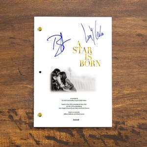 Pode incluir: Um roteiro branco com o título "A Star Is Born" em letras douradas, apresentando uma foto em preto e branco de um casal. O roteiro é assinado em tinta azul. O fundo é uma superfície de madeira.