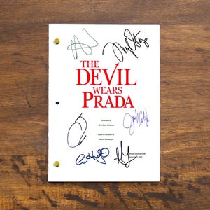 Pode incluir: Um roteiro branco com o título "O Diabo Veste Prada" em letras vermelhas. O roteiro é assinado por várias pessoas, incluindo o roteirista, o diretor e o produtor.