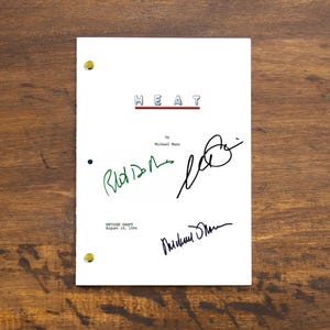 Pode incluir: Um roteiro branco intitulado "HEAT" com o título em vermelho e as assinaturas dos membros do elenco em tinta verde e preta. O roteiro é preso com fixadores de latão e repousa sobre uma superfície de madeira.