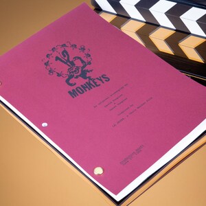 Puede incluir: Una cubierta de guion de película color burdeos con el título "12 Monkeys" impreso en negro. La cubierta tiene una claqueta en blanco y negro en el fondo.