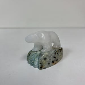 May include: A small, white polar bear sculpture carved from a translucent material. The bear is perched on a mottled, rectangular base with shades of green, gray, and brown. The sculpture is a decorative item.