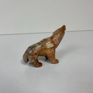 May include: A small, carved stone figurine of a wolf in a howling pose. The sculpture is primarily brown with mottled green and tan markings. The wolf is depicted with its head raised and mouth open, suggesting a howl.