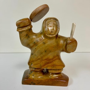 Könnte beinhalten: Eine handgeschnitzte Holzskulptur einer Person, die einen Kapuzenparka trägt und eine Trommel und ein Messer hält. Die Skulptur ist aus hellem braunem Holz mit dunkelbraunen Akzenten gefertigt.