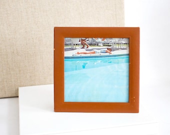 May include: A square, brown picture frame with a photograph of a person lying by a pool. The frame is made of a smooth, solid material. The photograph shows a person in light-colored clothing near a pool with blue water.