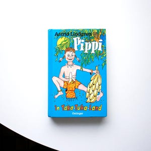 Può includere: Un libro con copertina rigida blu intitolato "Pippi in Taka-Tuka-Land" di Astrid Lindgren. La copertina presenta un'illustrazione di una ragazza con trecce rosse che tiene delle banane. La ragazza indossa un abito a tema banana.