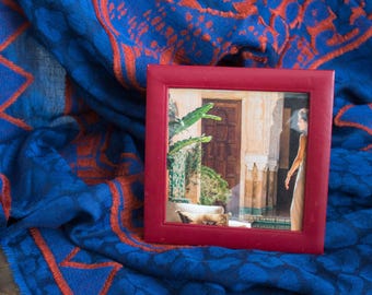 May include: A small, square, burgundy picture frame displaying a painting of a courtyard scene with a woman. The frame rests on a blue and orange patterned textile.