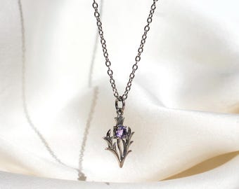 May include: A silver-toned pendant necklace featuring a stylized floral design with a central square-cut purple gemstone. The pendant hangs from a delicate chain, set against a white background.