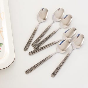Peut inclure: Ensemble de six cuillères en argent avec des manches gravés. Les cuillères, de forme ovale, sont disposées sur une surface blanche. Les manches présentent un motif floral détaillé.