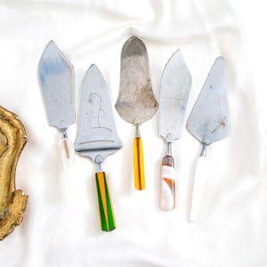 Op de afbeelding: Vijf vintage taartscheppen met zilveren bladen en diverse handvatontwerpen. De handvatten hebben witte, oranje, groene en patroonaccenten. De scheppen zijn gerangschikt op een wit, getextureerd oppervlak.