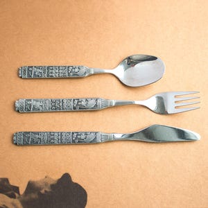 May include: A set of three silver-colored utensils: a spoon, fork, and knife. The handles feature a detailed, repeating pattern of figures and designs. The utensils are arranged on a light brown surface.