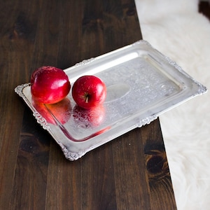 Könnte beinhalten: Ein rechteckiges silbernes Serviertablett mit verzierten Kanten hält zwei glänzende rote Äpfel. Das Tablett steht auf einer dunklen Holzoberfläche, mit einem weißen, flauschigen Teppich im Hintergrund. Das Tablett hat ein Blumenmuster.