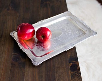 Può includere: Un vassoio rettangolare d'argento con bordi decorati contiene due mele rosse lucenti. Il vassoio è appoggiato su una superficie di legno scuro, con un tappeto bianco e soffice sullo sfondo. Il vassoio ha un motivo floreale.