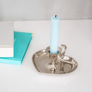 Può includere: Un portacandele color argento con base a forma di cuore e una piccola maniglia. Una candela azzurra è al centro. Accanto al portacandele ci sono due libri, uno bianco e uno turchese.