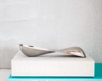 May include: A silver-colored, abstract sculpture with a curved, ribbon-like design. The sculpture is placed on top of a stack of books with the text "minimum" visible on the top book and "A LIST A DAY" on the bottom book.