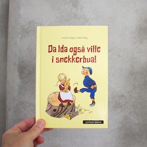 Può includere: Un libro per bambini giallo intitolato "Da Ida også ville i snekkerbua!" presenta illustrazioni di due bambini e un ceppo d'albero. La copertina è un disegno colorato in stile vintage. Il libro è tenuto da una mano.