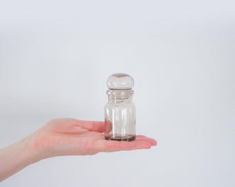 May include: A small, clear glass jar with a rounded lid. The jar is held in the palm of a hand against a white background. The jar has a cylindrical body and a small, round lid. The glass has a slight tint of brown.