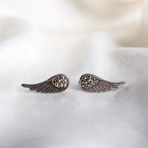 May include: A pair of silver-toned wing-shaped earrings. Each earring features a detailed feather design with a cluster of small, dark stones at the base. The earrings are displayed on a white, textured surface.