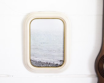 May include: An off-white rectangular picture frame with a gold inner edge. The frame displays a grayscale image of a seascape with a rocky shore. The frame is mounted on a white wall, with a section of a wooden decorative piece visible on the right.