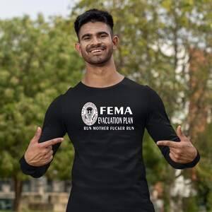 A man outdoors gesturing toward the FEMA evacuation plan graphic on his black long-sleeve shirt.