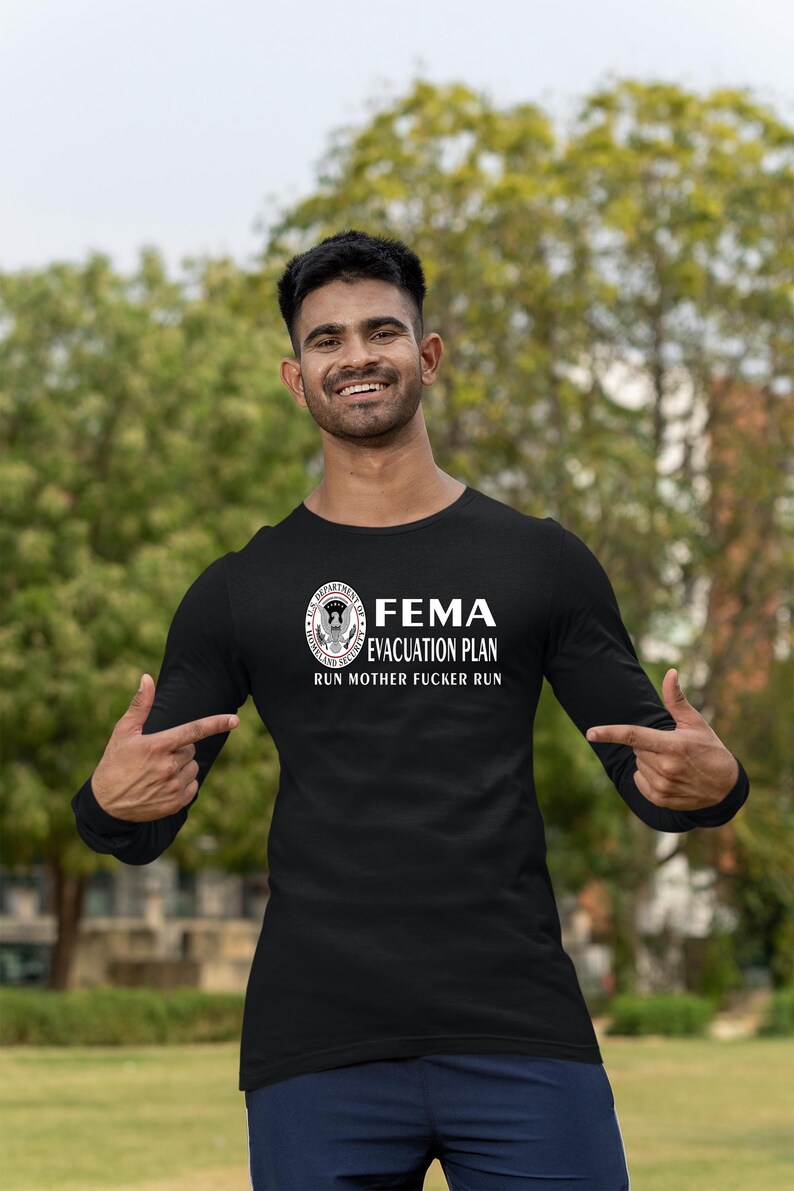 A man outdoors gesturing toward the FEMA evacuation plan graphic on his black long-sleeve shirt.