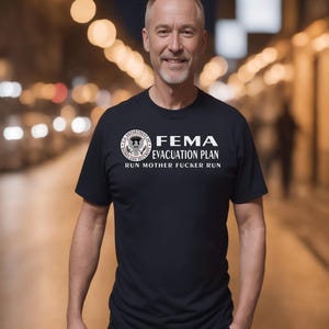 A mature man smiling outdoors in a black t-shirt displaying a humorous FEMA emergency design.